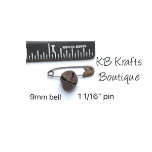 May include: A small, rusty-colored bell attached to a safety pin. The bell measures 9mm, and the pin is 1 1/16 inches long. The text "KB Krafts Boutique" is visible. A ruler is in the background.