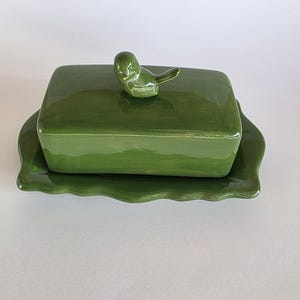Può includere: Una burriera in ceramica verde con una maniglia a forma di uccello sul coperchio. Il piatto rettangolare poggia su un vassoio smerlato abbinato. Il colore è un verde vibrante e uniforme.