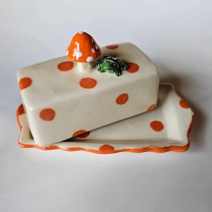 Puede incluir: Una mantequera de cerámica con tapa y bandeja a juego. El plato es de color blanquecino con lunares naranjas y una seta naranja decorativa y una rana verde en la parte superior. La bandeja tiene un borde naranja festoneado.