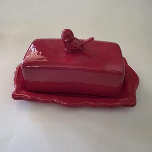 Puede incluir: Una mantequera de cerámica roja con tapa y una pequeña figura de pájaro. El plato se asienta sobre una bandeja a juego con un borde ondulado. La mantequera es de forma rectangular.
