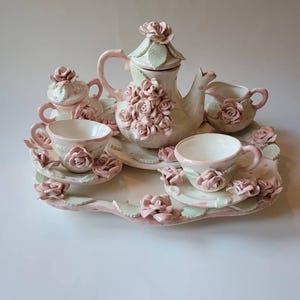 May include: A ceramic tea set with a tray, teapot, sugar bowl, creamer, and two teacups. The set is decorated with pink roses and green leaves. The tray measures approximately 12 inches in length.