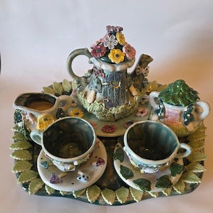 May include: A whimsical ceramic tea set featuring a teapot, creamer, sugar bowl, and two teacups with saucers. The teapot is adorned with colorful flowers and a fairy house design. The set is placed on a tray with leaf and floral details.