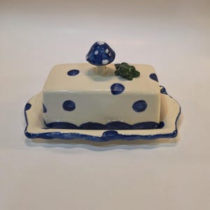 May include: A ceramic butter dish with a lid, featuring a cream-colored base with blue polka dots. The lid is topped with a blue mushroom and a small green turtle. The dish sits on a matching scalloped tray, with blue trim.