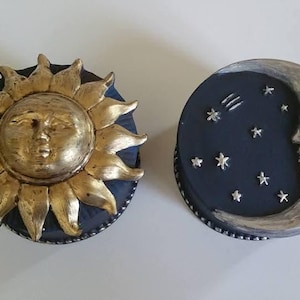 Puede incluir: Dos objetos decorativos con temática celestial. Uno es un sol dorado con rostro y rayos, sobre una base azul oscuro. El otro es una luna plateada con estrellas y una estrella fugaz sobre una base azul oscuro.
