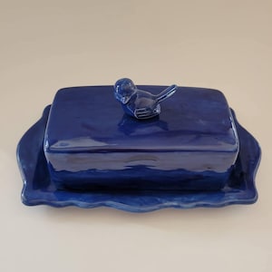 Könnte beinhalten: Ein kobaltblaues Keramik-Buttergericht mit passendem Deckel und Boden. Der Deckel ist rechteckig und hat einen vogelförmigen Griff. Der Boden hat einen gewellten Rand. Das Gericht ist zum Aufbewahren von Butter bestimmt.