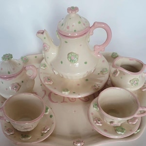 May include: A ceramic tea set with a teapot, sugar bowl, creamer, two cups, and saucers, all on a tray. The set is off-white with pink and green floral and polka dot accents. The tray has a pink border.