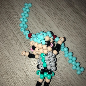 May include: A handmade figurine crafted from colorful beads. The figure features a light blue hairstyle, black and teal clothing, and a pink face. The arms and legs are black and tan. The figure is on a wooden surface.