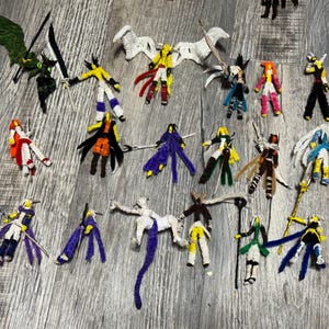 May include: A collection of handmade pipe cleaner figures, each depicting a unique character with swords, wings, and various colored outfits. The figures are arranged on a gray wooden surface, showcasing a variety of designs and poses.