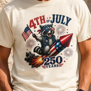 Puede incluir: Una camiseta de color crema con un gráfico de un mapache con bandana y gafas de sol, montado en un cohete de fuegos artificiales. El diseño incluye una bandera estadounidense, fuegos artificiales y el texto 4TH OF JULY 250 YEARS.