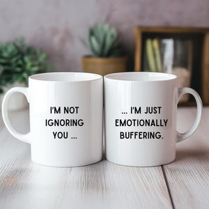 May include: Two white ceramic mugs with black text. One mug reads "I'M NOT IGNORING YOU ..." and the other reads "... I'M JUST EMOTIONALLY BUFFERING."