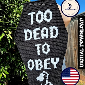 May include: A black coffin-shaped crochet wall hanging featuring the white text TOO DEAD TO OBEY and a white graphic of a wilted flower at the bottom. The piece includes the small text @thebloodstitch near the top edge and hangs from a black cord.