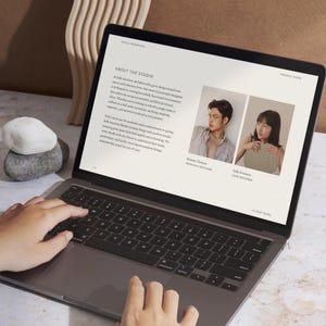 Puede incluir: Un ordenador port&aacute;til muestra un sitio web con texto y dos fotos de perfil. La pantalla muestra las palabras "About The Studio" y los nombres de dos personas. Se ven manos tecleando en el teclado. Piedras decorativas y una escultura de madera en el fondo.