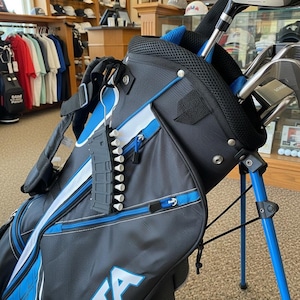 May include: A black and blue golf bag with a stand, featuring the word SATA printed in large white letters. The bag includes a black plastic golf tee holder shaped like a magazine, holding several white golf tees, attached to the side.
