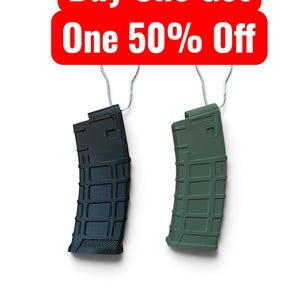 May include: Two polymer firearm magazines, one black and one olive green, each featuring a textured grid pattern and a metal wire hanger. Above the magazines is a red rectangular sign with white text that reads Buy One Get One 50% Off.