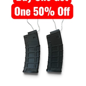 May include: Two black polymer rifle magazines with a textured grid pattern on the exterior surface. Each magazine features a metal wire hanger attached to the top. Above the magazines, a red rectangular sign displays the text Buy One Get One 50% Off.