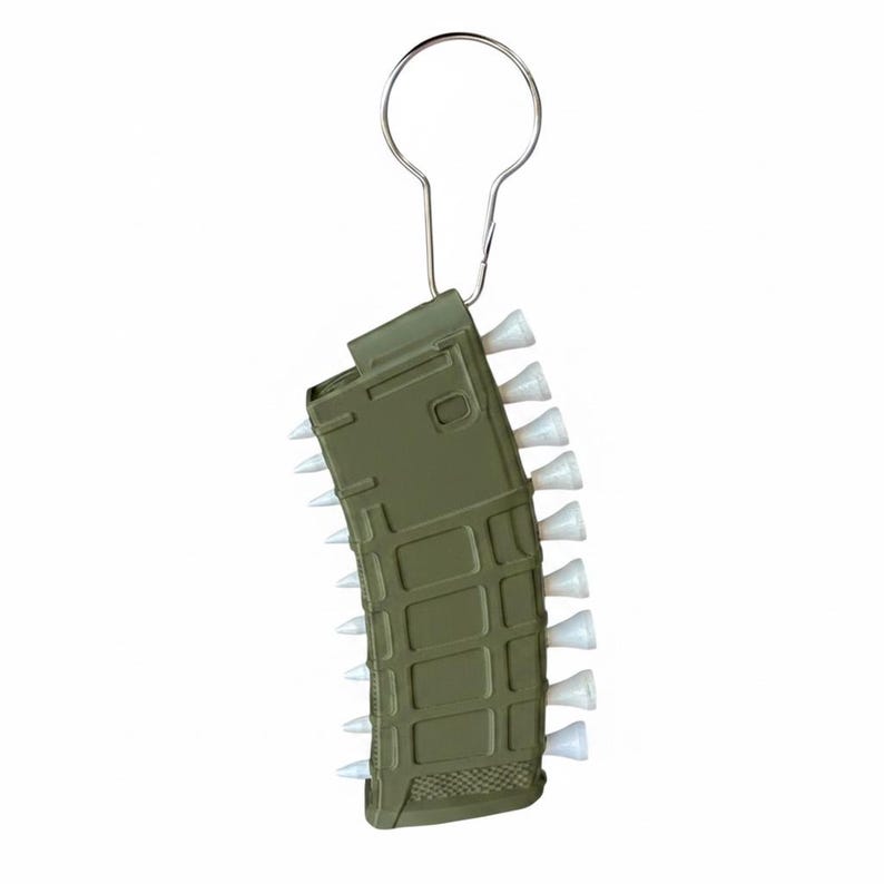 May include: An olive green rifle magazine shaped keyring featuring a series of small white golf tees inserted along the side edges. The object is attached to a silver metal split ring at the top, designed for holding keys or other small items.