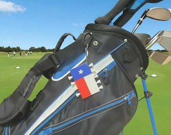 May include: A black and blue golf bag with a rectangular tag featuring the Texas state flag design. The tag is attached to the bag with a black carabiner and holds several white golf tees along its vertical edges.