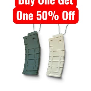 May include: Two rifle magazines, one olive green and one tan, hang from metal hooks. Above them, a red rectangular sign with rounded corners displays the white text: Buy One Get One 50% Off. The magazines feature a grid-like textured surface.
