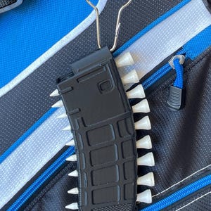 May include: A black plastic magazine-shaped golf tee holder featuring a grid-like textured surface. The holder is attached to a silver metal carabiner and contains multiple white golf tees inserted along both of its vertical sides.
