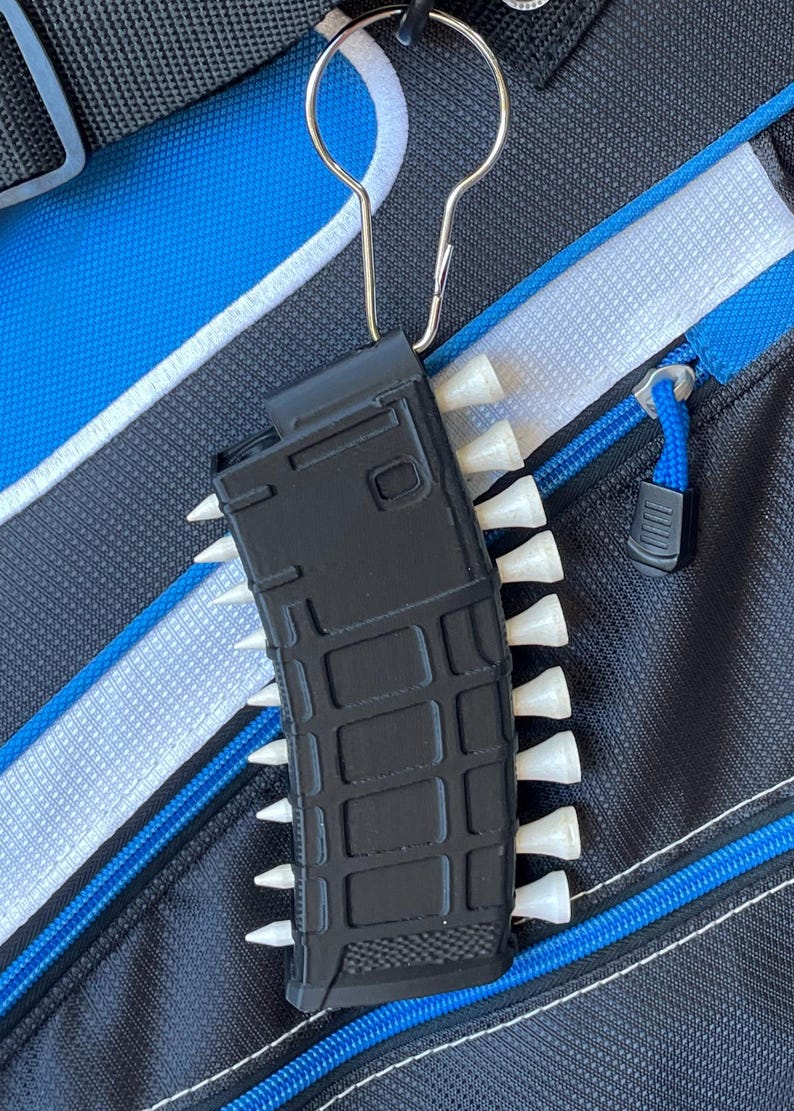 May include: A black plastic magazine-shaped golf tee holder featuring a grid-like textured surface. The holder is attached to a silver metal carabiner and contains multiple white golf tees inserted along both of its vertical sides.