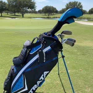 May include: A black and blue Strata golf bag with a stand, featuring white accents and the brand name printed in white. The bag contains several golf clubs with headcovers, including a driver with a blue cover, and a small black accessory.
