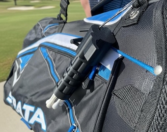 May include: A black plastic golf tee holder attached to a black and blue golf bag. The holder features a cylindrical design with vertical ridges and holds several white golf tees. The golf bag has white trim and the partial text STRATA visible.