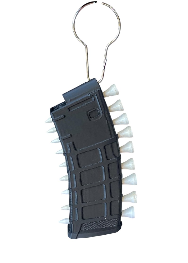 May include: A black, rectangular, textured magazine-shaped keyring featuring a series of small, white, cone-shaped golf tees inserted along both sides. The object is attached to a silver-coloured metal keyring loop at the top.