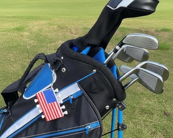 May include: A black and blue golf bag with a stand, containing several metal golf clubs with black headcovers. A small rectangular tag featuring the American flag is attached to the side of the bag, which displays the partial white text STRATA.