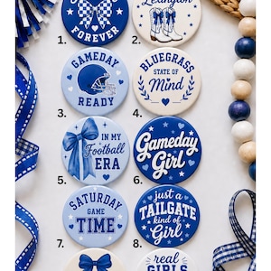 May include: A collection of ten round buttons featuring blue and white designs with football-themed text and graphics. The buttons include phrases like Football Forever, Game Days in Lexington, Gameday Ready, Bluegrass State of Mind, In My Football Era, Gameday Girl, Saturday Game Time, Just a Tailgate Kind of Girl, Saturday Sweetheart, and Real Girls Love Football.