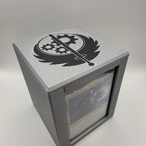 May include: A gray card storage box with a silver top featuring a black graphic design of a gear, sword, and wing-like shapes. The box has a clear window displaying a card with the text "Liberty Prime, Recharged".
