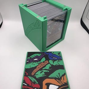 May include: Green card storage box with a lid featuring a Teenage Mutant Ninja Turtles design. The box is filled with cards and has the word "Cowabunga!" printed on the side. The lid has a colorful graphic of the turtles.