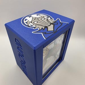 May include: A blue, cube-shaped card storage box with a character graphic on the top. The box has a clear window on the front and the word "MOTORIND" is printed on the side. The character graphic is white and gray.