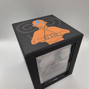 May include: Black display case featuring a card with the text "Avatar Aang." The top of the case has an orange and blue graphic of Avatar Aang. The case is designed to protect and showcase collectible cards.