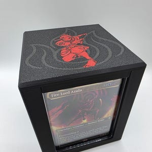 May include: Black card storage box with a red illustration of a character on the top. The box has a clear window displaying a Magic: The Gathering card with the text "Fire Lord Azula". The box is designed to hold trading cards.