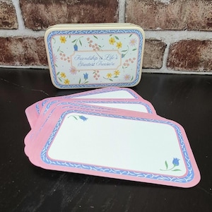 May include: A rectangular, off-white tin box and several pink name cards. The box is decorated with a floral pattern and the words "Friendship is Life's Greatest Treasure." The name cards have a pink border with a blue floral design.