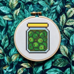 以下が含まれることがあります： 木製の刺繍枠にセットされた、赤いアクセントの緑色のオリーブの瓶のクロスステッチ刺繍。瓶には黄色の蓋があり、緑色のオリーブが詰まっています。背景は、葉の模様の濃いティール色の生地です。