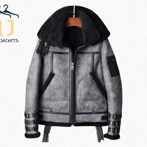 May include: A grey shearling jacket with a black shearling collar and lining. The jacket has a front zip, multiple pockets, and buckle details on the sleeves. The text "NOIR JACKETS" is visible in the upper left corner.