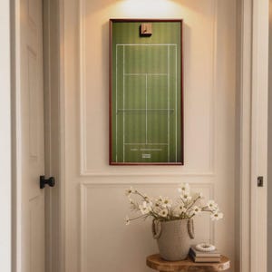 May include: A framed tennis court print hangs on a white wall, above a small wooden stool with a woven basket of white flowers. The print has a green court with white lines and a brown frame. The text "Tennis Frame" is visible.