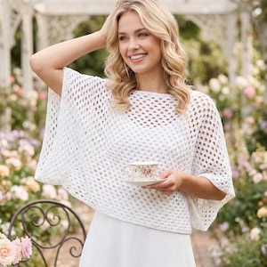 May include: A woman wearing a white, open-weave crochet poncho. She is holding a floral teacup and saucer. The background features a garden with flowers and a white arbor. The poncho has a square neckline and short sleeves.