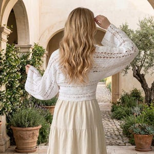 May include: A woman wearing a white, open-weave crochet top with long, flared sleeves and a cream-coloured skirt. The outfit is set against an outdoor courtyard with plants and stone arches.