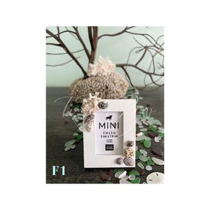 May include: A white mini picture frame, 2 in x 3 in (5 cm x 7.6 cm), decorated with seashells and small stones. The frame has the word "MINI" and a dog silhouette printed on it. The frame is surrounded by sea glass and other decorative elements.