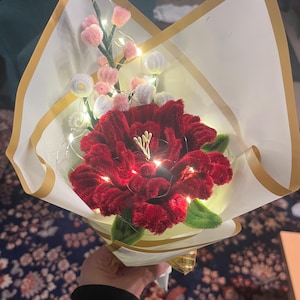 Könnte beinhalten: Ein handgefertigter Blumenstrauß mit einer großen, plüschigen roten Blume als Mittelpunkt, umgeben von kleineren rosa und weißen Pomponblumen. Der Strauß ist in weißes Papier mit goldenem Rand gewickelt und mit Lichterketten beleuchtet.