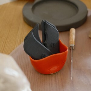 May include: An orange ceramic bowl contains several dark grey pottery tools. A small metal knife with a wooden handle sits beside the bowl. In the background are clay pieces and a grey ceramic plate on a wooden table.