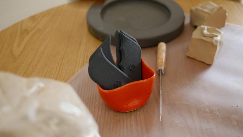 May include: An orange ceramic bowl contains several dark grey pottery tools. A small metal knife with a wooden handle sits beside the bowl. In the background are clay pieces and a grey ceramic plate on a wooden table.