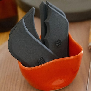 Puede incluir: Un juego de cuatro utensilios de cocina curvos de color gris oscuro con una superficie texturizada, colocados en un pequeño cuenco de cerámica naranja brillante. Cada utensilio tiene una forma única y un pequeño diseño circular. El cuenco está sobre una superficie de madera clara.