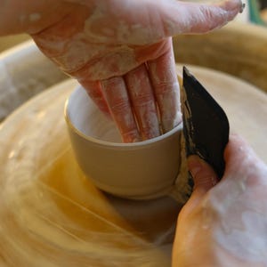 May include: A potter's hands shaping a small, beige ceramic cup on a pottery wheel. The hands are covered in wet clay, using a black tool to refine the cup's form. The wheel and surrounding area are also covered in clay.