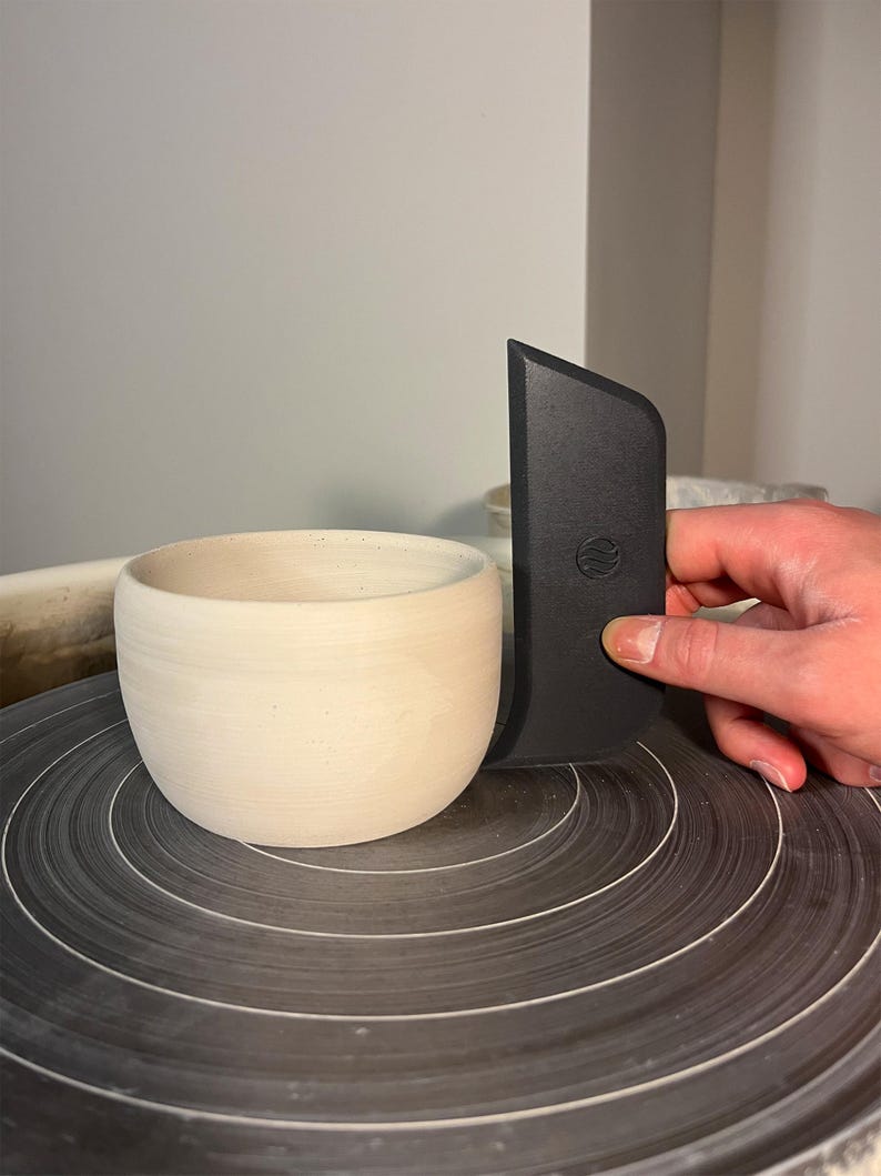 May include: A cream-coloured ceramic bowl sits on a pottery wheel. A black trimming tool is held next to the bowl. The bowl has a rounded shape and a smooth, unglazed surface. The pottery wheel is dark grey with concentric circles.