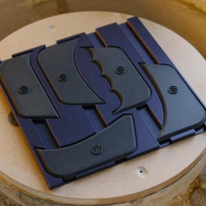 Könnte beinhalten: Ein Satz dunkelgrauer Töpferwerkzeuge, jedes mit einer einzigartigen Form und einem kleinen Emblem, angeordnet auf einer dunkelblauen Basis. Diese Werkzeuge dienen zum Formen und Glätten von Ton. Das Set wird auf einer Töpferscheibe präsentiert.