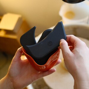 May include: A person holding a set of pottery tools. The tools include a dark grey rib tool and a black rib tool, nested inside a small, orange ceramic bowl. The tools are used for shaping and smoothing clay.