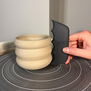 Op de afbeelding: Een beige keramische pot met een geribbeld ontwerp. Een zwart pottenbakkersgereedschap wordt naast de pot gehouden. De pot staat op een zwart pottenbakkerswiel met concentrische witte cirkels.